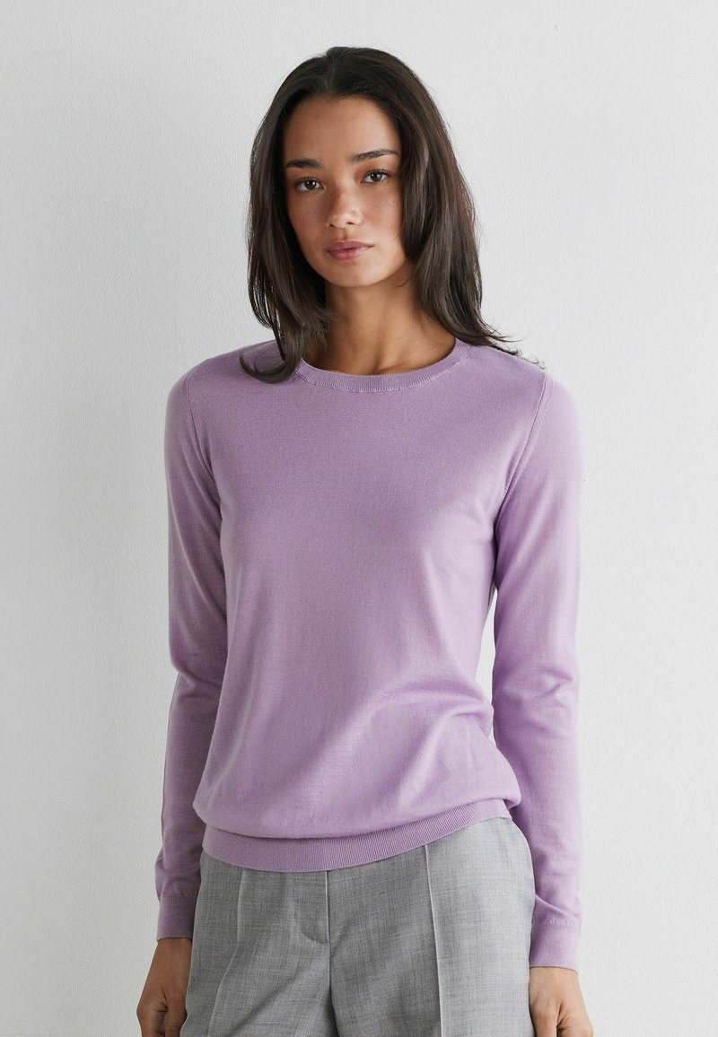 Kvinde med langt mørkt hår iført en lavendel farvet trøje med lange ærmer og grå højttaljede bukser, stående mod en ensfarvet hvid væg.