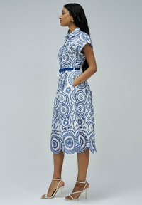 Vestido de estampado floral en azul y blanco, con mangas cortas, cintura con cinturón y bolsillos. El material parece ligero y tiene un dobladillo en forma de ondas.