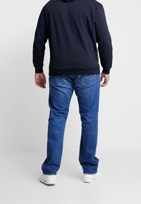 Blue denim jeans with a straight cut, featuring back pockets and stitching details. Worn with a dark hooded sweatshirt and white sneakers.