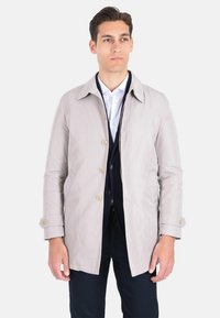 Impermeabile beige chiaro con finitura testurizzata, con colletto classico, bottoni frontali e polsini regolabili, abbinato a pantaloni blu navy.