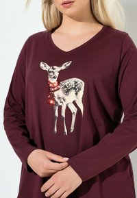Bordeaux-punaiset pitkähihainen paita, jossa on graafinen kuva hirvestä, joka käyttää paljettihuivia. V-kaula-aukko, pehmeä kangas ja rento leikkaus.