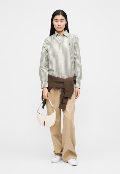 Pruhovaná košeľa na zapínanie na gombíky v zelenej a bielej farbe, hnedý sveter omotaný okolo pása, béžové nohavice, drží bielu tašku, biele tenisky.