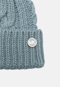 Beanie lavorato a maglia in morbido filato blu con un design a coste. Presenta una spilla rotonda in argento con "MK" sul davanti.