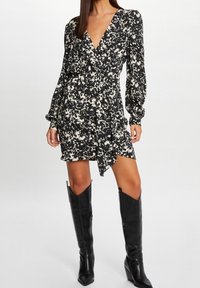 Robe portefeuille fleurie noire et crème avec manches longues bouffantes, col en V, taille nouée et ourlet court, associée à des bottes en cuir noires montantes jusqu'aux genoux.