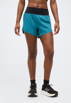 Αθλητικό άτομο που φοράει πετρόλ σορτς τρεξίματος με μαύρη ζώνη μέσης, μαύρες κάλτσες και μαύρα παπούτσια τρεξίματος New Balance σε απλό φόντο.