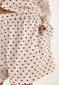 Pantaloni da pigiama in cotone con elastico in vita, caratterizzati da una base bianca e motivi a cuori rossi. Il tessuto ha un aspetto strutturato.