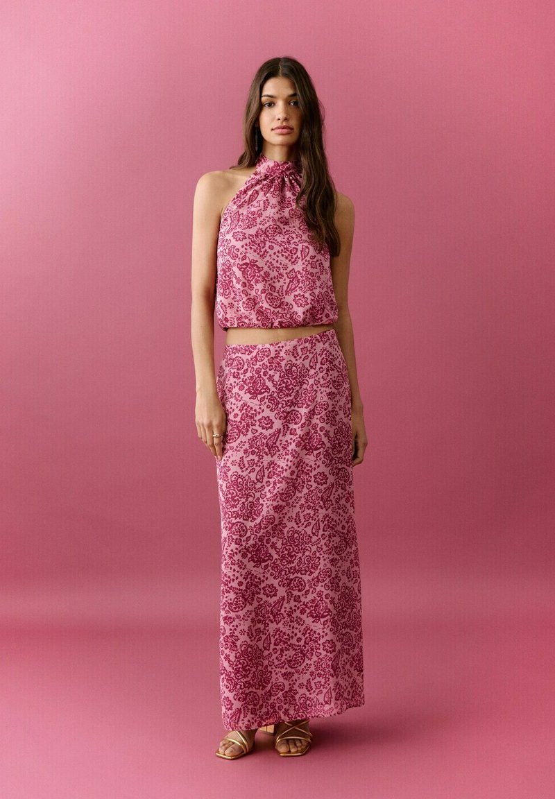 Kvinde med langt mørkt hår iført ærmeløs lyserød blomster-haltertop og matchende lang nederdel, stående mod ensfarvet lyserød baggrund.