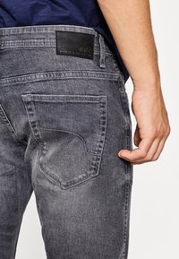 Primer plano de la parte inferior de una persona que lleva unos vaqueros grises desgastados con un bolsillo trasero y una etiqueta de marca de cuero negro, con la mano descansando al lado.