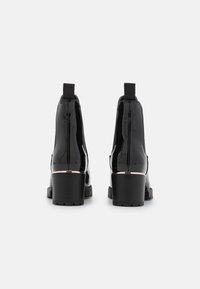 Botas de tobillo de charol negro con paneles laterales elásticos, tacones cuadrados y una banda de acento dorado alrededor del tacón. Suela texturizada para mayor agarre.
