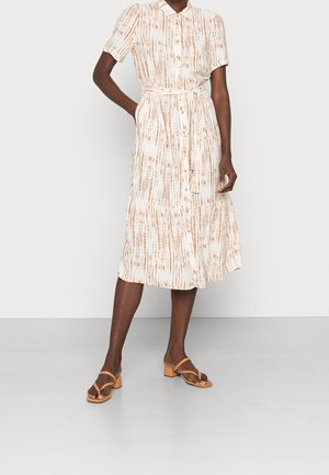 Frau trägt ein beigefarbenes und braun gestreiftes Midikleid mit kurzen Ärmeln und Bindegürtel an der Taille, kombiniert mit hellbraunen Sandaletten mit Absatz.