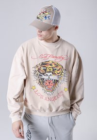 Felpa beige oversize con grafica tigre e testo rosso; pantaloni della tuta grigi con lacci. Cappellino trucker beige e bianco con design di aquila.