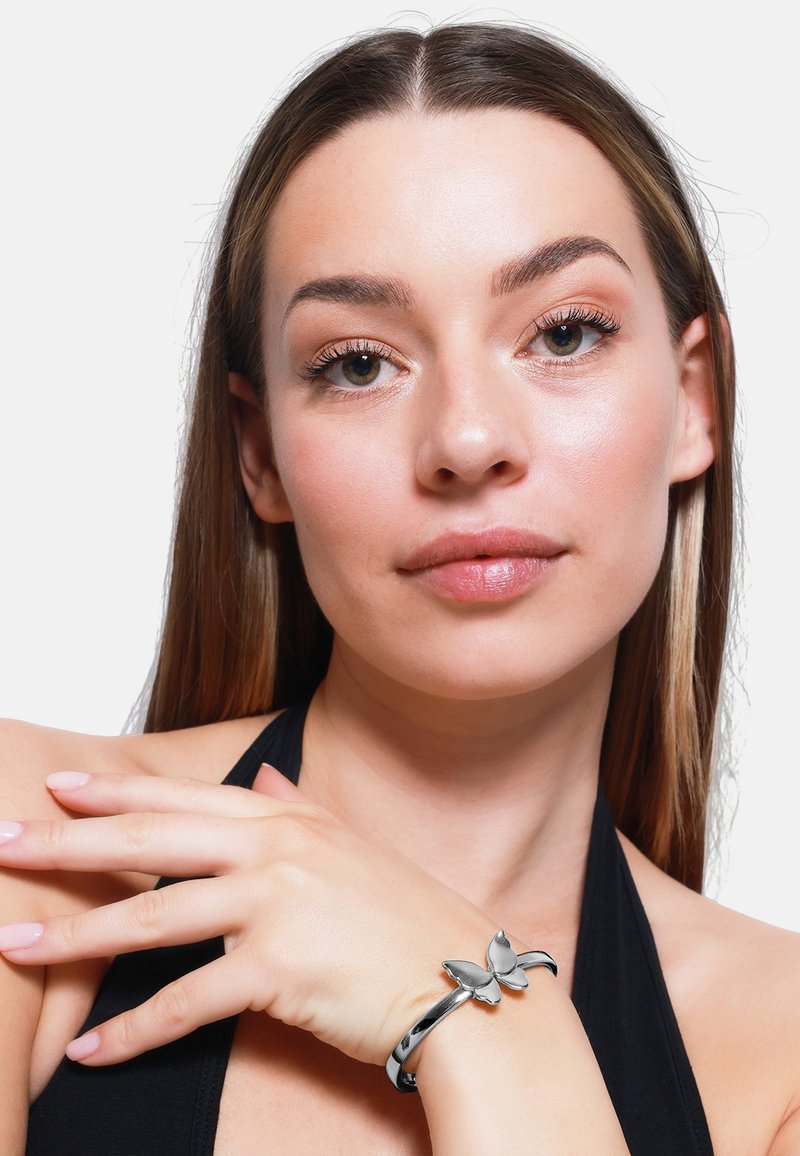 Bracciale in argento con design a farfalla, superficie metallica liscia e finitura lucida, indossato su un polso su uno sfondo semplice.