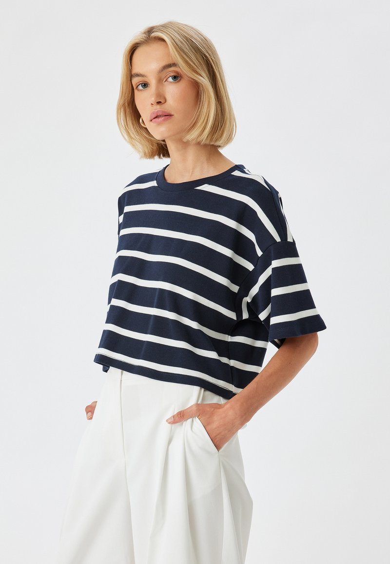 Camiseta corta de rayas azul marino y blanca, con mangas cortas y escote redondo, fabricada en suave algodón, combinada con pantalones blancos holgados, con las manos en los bolsillos.