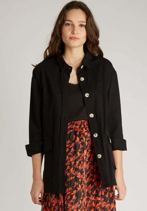 Veste noire avec un col, boutons argentés et manches retroussées, associée à une jupe à motifs floraux rouges et noirs. Tissu léger.