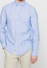 Chemise à rayures bleu et blanc, boutonnée, en tissu léger ; dotée d'un col pointu et de manches longues, avec un ourlet arrondi.