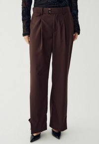 Smeđe, plisirane hlače s visokim strukom, opremljene s dvije dugmadi, prednjim džepovima i ravnim krojem. Glatka tekstura i krojena izrada.