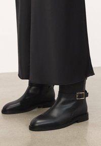 Black leather ankle boots with a smooth texture, a rounded toe, and a strap with a square gold buckle. Flat sole and simple design.