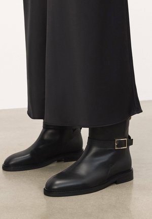 Black leather ankle boots with a smooth texture, a rounded toe, and a strap with a square gold buckle. Flat sole and simple design.