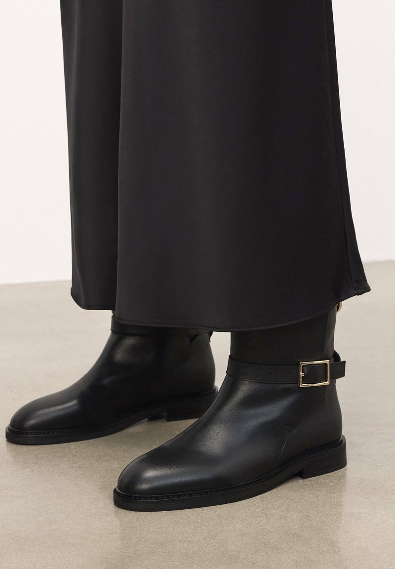 Black leather ankle boots with a smooth texture, a rounded toe, and a strap with a square gold buckle. Flat sole and simple design.