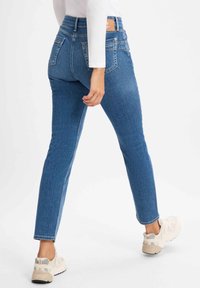 Schmale blaue Jeans mit mittlerer Taille, Gesäßtaschen und dezentem Verblassen. Kombiniert mit hellen Sneakern.