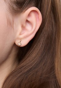 Złote kolczyki w kształcie serca z małym, przezroczystym kamieniem szlachetnym. Posiadają cienką, drucianą konstrukcję i są pewnie osadzone w płatku ucha.