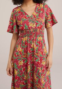 Czerwona sukienka w kwiaty z dekoltem w kształcie litery V, krótkimi rękawami, marszczonym pasem oraz zielonymi i żółtymi wzorami kwiatowymi. Wykonana z lekkiego materiału.