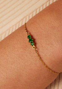 Gouden kettingarmband met drie groene edelstenen, rond geslepen, bevestigd in een decoratieve klauwbevestiging op een huidkleurige pols.