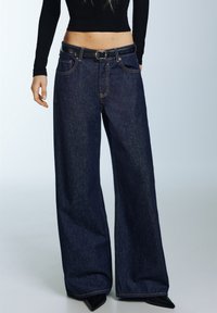 Traperice širokih nogavica tamnoplave boje, s visokim strukom, rupicama za remen i kontrastnim šavovima; u kombinaciji s crnim topom i cipelama s vrhovima ušiljenim.