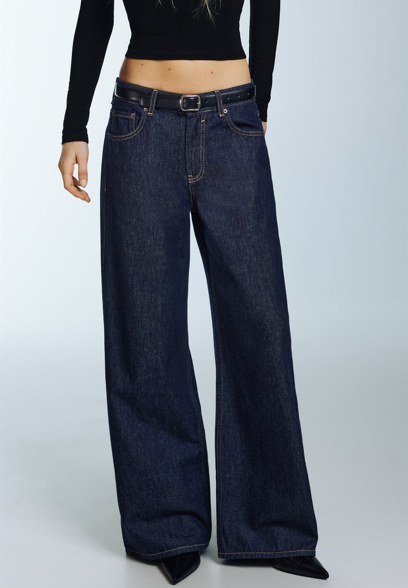 Traperice širokih nogavica tamnoplave boje, s visokim strukom, rupicama za remen i kontrastnim šavovima; u kombinaciji s crnim topom i cipelama s vrhovima ušiljenim.