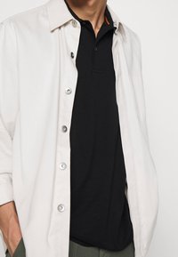 Ljust beige knapp-up jacka över en svart polo shirt. Jackan har silverfärgade knappar och en enkel krage, med en slät textur.