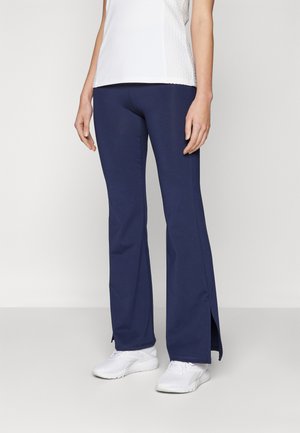 Navy blue flared pants with side slits made of a stretchy, breathable material. Worn with white athletic shoes and a white, textured top.