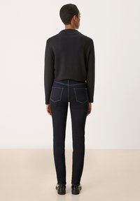 Jeans skinny bleu foncé avec des coutures jaunes contrastantes. Porté avec un haut noir à manches longues, mettant en valeur une silhouette ajustée et une texture lisse.