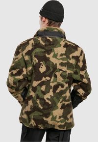 Maastokuvioinen fleece-takki, jonka pinta on teksturoitu sävyissä vihreä, ruskea ja beige; siinä on kontrastiväriset mustat kaulus ja hihansuut.