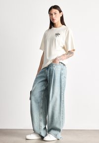 Krémové bavlnené tričko s grafickou potlačou, kombinované so svetlomodrými širokými džínsami a bielymi teniskami, ktoré zdôrazňujú pohodlný strih a ležérny štýl.