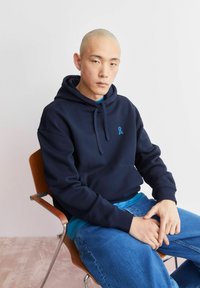 Donkerblauwe hoodie met een blauw logo, gemaakt van zachte stof. Draagt over een lichtblauw shirt, gecombineerd met een spijkerbroek.