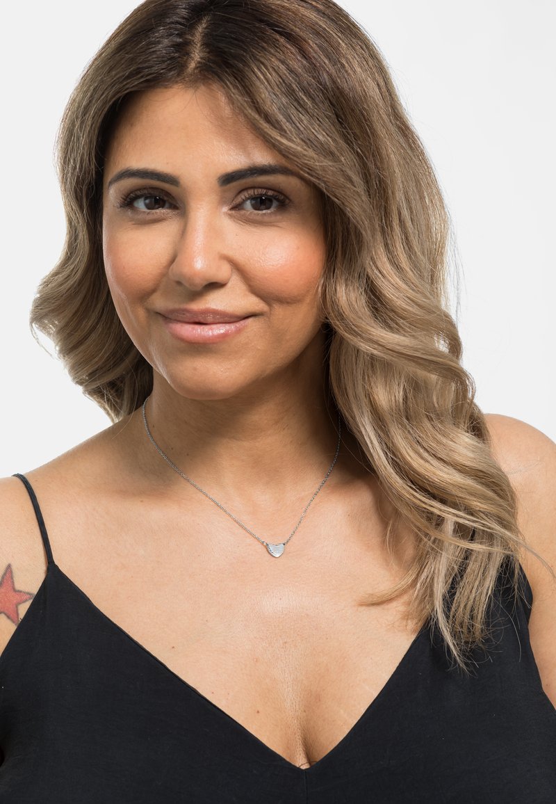 Collier en argent avec un pendentif en forme de cœur, porté avec un haut noir à col en V. Le pendentif a une surface texturée et une fine chaîne.