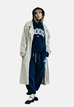 Navy Kapuzenpullover mit "GOOD" Druck, kombiniert mit passenden Jogginghosen, darüber getragen mit einem hellbeigen Trenchcoat. Schwarze Ballerinas und weiße Socken.