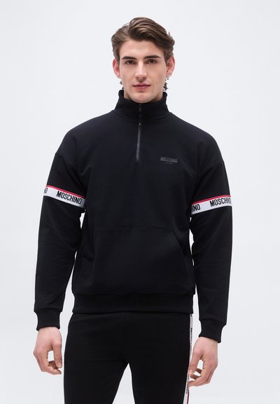 Jeune homme portant un pull Moschino noir avec une demi-fermeture éclair et des bandes blanches et rouges de marque sur les manches, debout devant un fond clair uni.