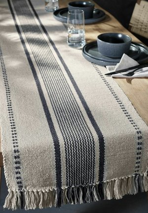 Beżowy tkany bieżnik na stół z czarnymi paskami i frędzlami na drewnianym stole nakrytym czarnymi talerzami, miskami, pałeczkami i szklankami wody.