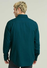 Camicia a maniche lunghe color turchese con colletto, polsini abbottonati e orlo dritto. Il tessuto appare liscio con una leggera lucentezza. Visibile la vista posteriore.