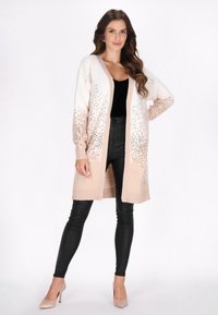 Stickad cardigan i kremfärg och blush-gradient, med detaljer vid nederkanten. Långa ärmar och öppen framsida, kombinerad med svarta byxor.