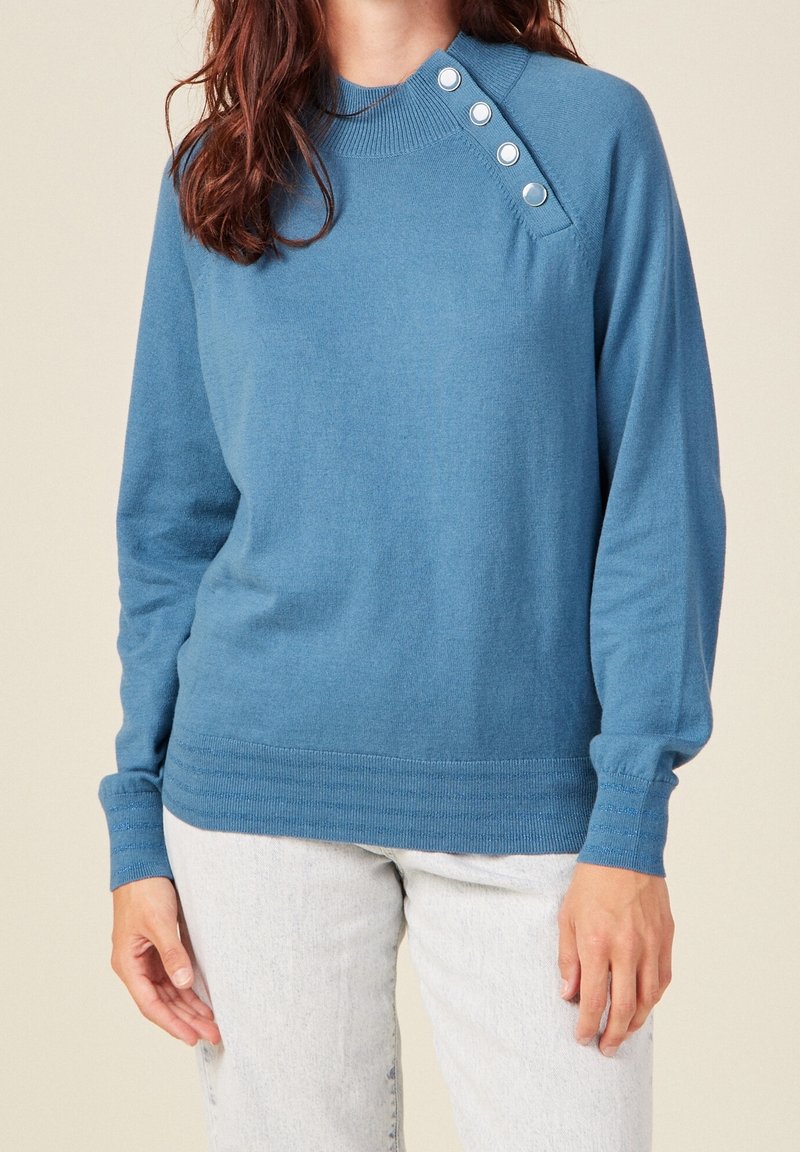 Pull bleu avec un col haut, comportant quatre boutons à l'épaule. Texture douce et poignets côtelés. Associé à un jean clair.