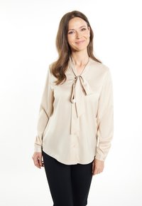 Beige samettipaita, jossa on solmuke-kaulus, pitkät hihat ja napit. Pehmeä rakenne, joka laskeutuu kauniisti.