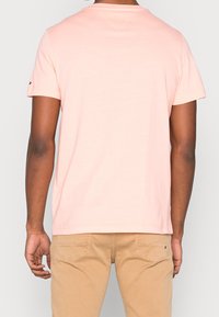 Camiseta de algodón color rosa claro con mangas cortas, cuello redondo y un sutil detalle de logo en la manga izquierda.