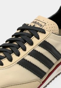 Μπεζ αθλητικό παπούτσι Adidas με μαύρα κορδόνια, τρεις μαύρες ρίγες στο πλάι και κόκκινη λεπτομέρεια πάνω από την σόλα.