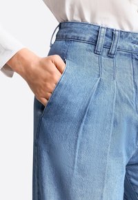 Mano en el bolsillo delantero de unos jeans de tiro alto de mezclilla azul claro, combinados con una camisa blanca de manga larga.
