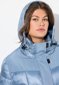 Chaqueta acolchada de color azul claro con capucha, con un diseño acolchado, cuello alto y detalles en negro, fabricada con material brillante y suave.