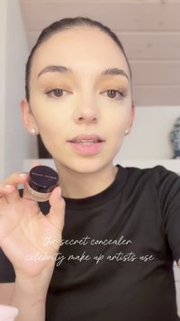 Piccola jar di vetro con un coperchio nero, contenente correttore beige. La persona la tiene sollevata, indossando una maglietta nera, con un trucco sobrio.