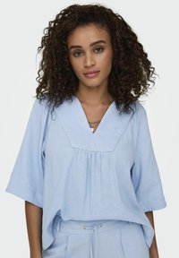 Top azzurro chiaro a maniche corte con scollo a V, design con pieghe sul davanti, realizzato in un tessuto testurizzato. Taglio semplice e casual, vestibilità comoda.
