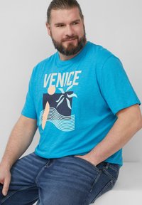 Behaarde man met vastgebonden haar, gekleed in een felblauw "VENICE" T-shirt met een abstracte palm- en golfontwerp, zittend met één hand in zijn zak.